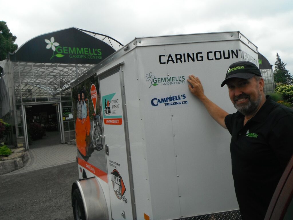 Photo of Rob Gemmell of Gemmell's Garden Centre pointing to his logo on the CWALC Trishaw trailer.